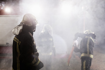 Team of professional firefighters with water hose in front of a firetruck with smoke in the air.