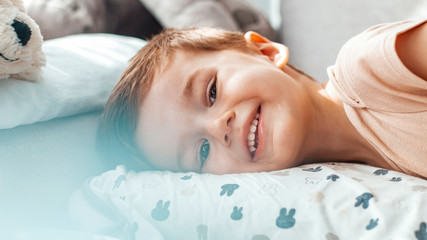 Obraz na płótnie Canvas Portrait of an adorable little boy smiling while lying down on a bed at home. Cute happy laughing boy. Handsome boy with toothy smile looking at camera. Lovely child lying on front in bed