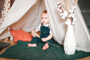 Sweet 1 year old little baby boy playing in a teepee game tent in a children's room decorated in...