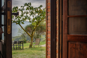 Opened wooden door of old brick farmer house presenting outstanding view of Toscany hills with single tree and table wit chairs around on sunless autumn day in Italy. Vacation in wine region.