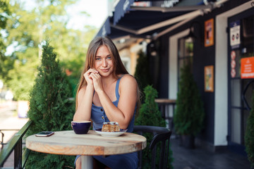 Cute cute girl smiles over a cup of tea. Stylish blue top with a skirt. Green thujas in a cafe.