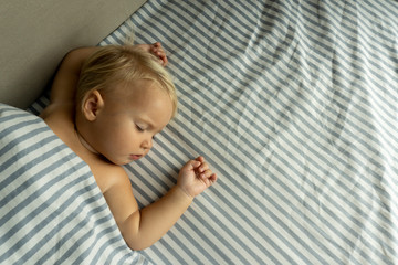 One year cute sweet blond child sleeps, raising hands up, on white and blue sheets. Top view, good background, copy space.