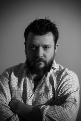 Black and white studio portrait of a brutal bearded man with a serious face in a light shirt holds his hands folded over his chest