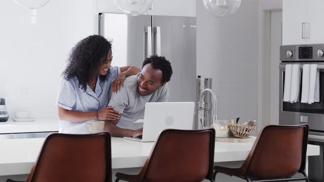 Couple Wearing Pyjamas Standing In Kitchen Working From Home On Laptop