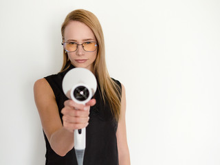Fototapeta premium Confident blonde female hairdresser in glasses and black apron standing against white wall in trendy barbershop salon holding professional hair dryer