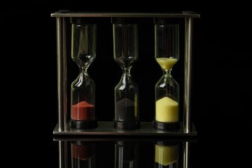 Fototapeta premium Three hourglasses with colorful sand stand on a black glossy glass, in which their reflection is visible.