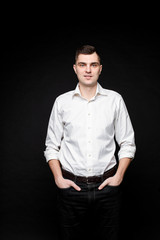 Tall cauasian male with short dark hair, pretty face in white shirt, black tousers keeps hands in his pockets