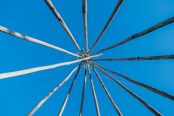 Naklejka na ściany i meble Native American wooden frame from a teepee tent temporary dwelling. Indigenous historical tipi shelter against blue sky and copy space background 