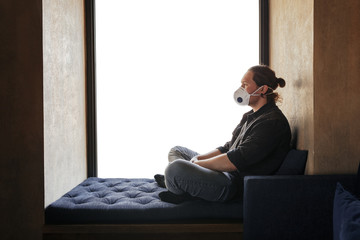 Fototapeta premium A lonely guy in self-defense with a protective medical mask is sitting by a big window looking at the world. The concept of isolation. The guy with the long hair, the hipster
