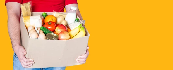 Cercles muraux Manger Un livreur d& 39 épicerie en uniforme rouge tient une boîte en carton avec des légumes frais, des fruits et d& 39 autres aliments sur fond orange. Livraison express de nourriture, concept de don.