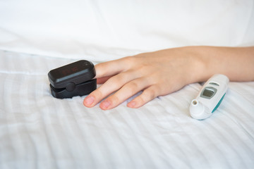 Woman's hand resting on bed with digital thermometer and wearing pulse oxymeter on finger