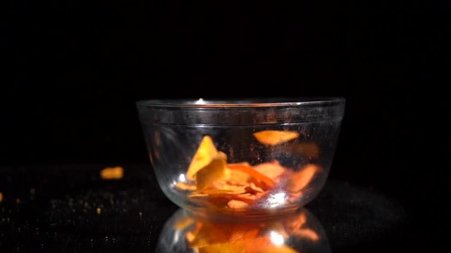 This slow motion video shows delicious cheese flavored tortilla chip snacks falling against a black background as they are poured into a clear glass bowl for serving.