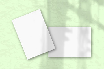 2 sheets of white textured paper against a gray wall. Mockup with an overlay of plant shadow Natural light casts shadows from the window. Flat lay, top view