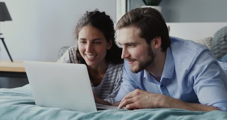 Happy millennial man and woman using laptop computer at home watching online show lying in bed together. Young adult couple looking at notebook screen making video call chatting in bedroom at home. - Powered by Adobe
