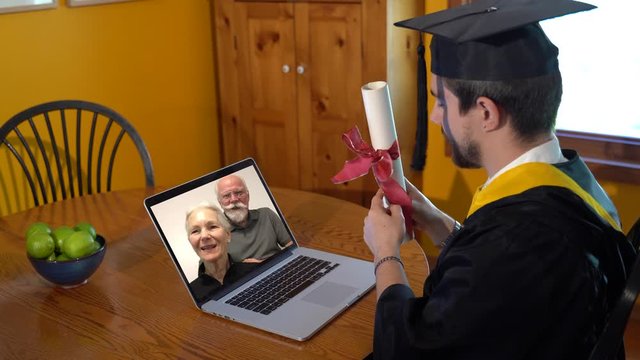 Side view of young man in graduation costume holding diploma and talking to grandparents online on a laptop.