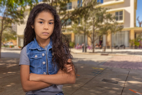 A young girl stands firm with her arms crossed, looking at you hard. She stands her ground downtown under a tree across the street from a sunlit outdoor cafe.
