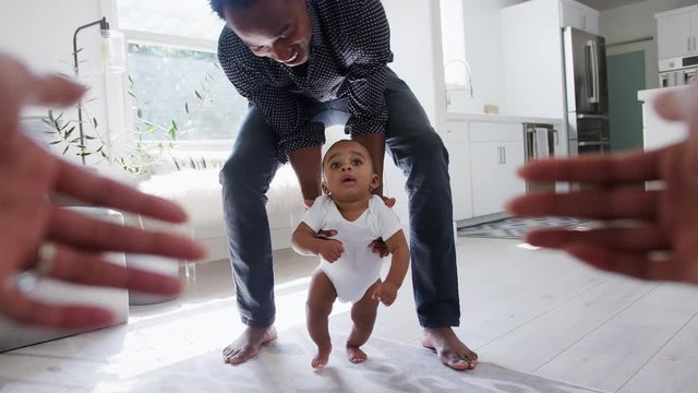 Point Of View Shot Of Parents Encouraging Smiling Baby Son To Take First Steps And Walk At Home