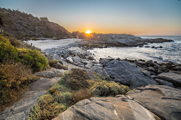 An amazing view of the sunset over the water in the Chilean coast. An idyllic beach scenery with the sunlight illuminating the rocks with orange tones and the sea in the background under a moody sky
