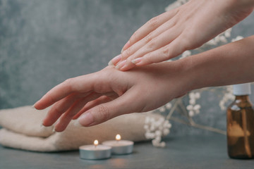 Obraz na płótnie Canvas Beautiful female hands care for the skin, apply cream and oils. Light fluffy beige towels with spa paraphernalia, candles and flowers on a dark gray background