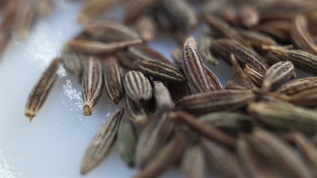 Cumin spices are scattered on a white background in extreme close-up. The camera hovers over the subject, smooth movement, real time, low depth of field