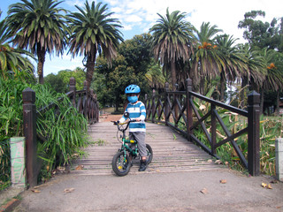 A young boy riding a bicycle and helmet on a bridge in a green park on a sunny day. Kid on a park bridge outside wearing a medical mask for the Coronavirus pandemic. Protection for the covis-19.
