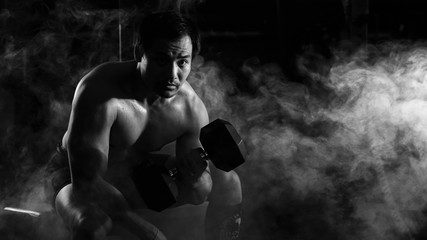 Fototapeta na wymiar asian athletic strong man sitting and having weight lifting with dumbbells with one arm in gym and fitness center with smoke and dark background in black and white