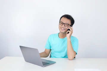 Young Asian Man with happy face is talking on a mobile phone with laptop on the table. Indonesian man wearing blue shirt.
