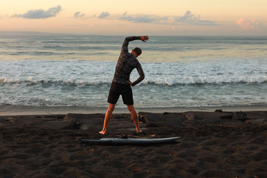 Surfing. Stretching Surfer With Surfboard On Sandy Beach At Sunrise In Bali. Man In Wetsuit Going To Surf In Ocean. Beautiful Sea And Sky Background.
