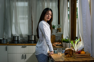 Stressed young beautiful Asian woman standing in the kitchen during quarantine. Loneliness sad girl living alone at home and thinking of problem. Mental health, unemployed and social issue concept.
