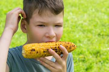 a boy outdoors on a background of green grass eats yellow corn and thinks of something, in front there is a place for inscription