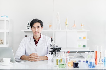 A male scientist with black hair wearing a white coat sitting with a laptop and a microscope in a laboratory with test tubes with his hand on table pose looking at the camera.