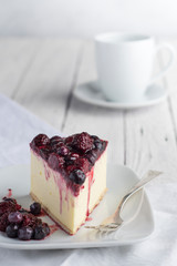 porción de pastel cheesecake con frutos rojos sobre un plato blanco, fondo blanco con una taza de café.