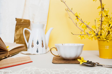 Fototapeta na wymiar white porcelain mug with and silver spoon with tea granules and yellow forsythia on light table, against the background of a white teapot, old books and a bouquet of spring flowers in a small bucket