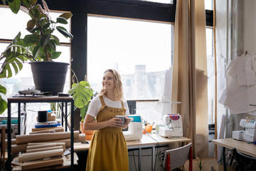 Happy female dressmaker resting at her workplace, drinking coffee.