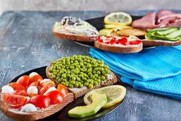 Assorted open-faced sandwiches, sandwiches with slices of sourdough with various fillings on a plate - cucumbers, peas, cherry tomatoes, mozzarella, sardines, bell peppers, pastramas, lemon