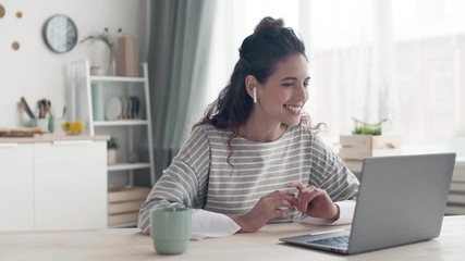 Medium shot of young cheerful woman sitting kitchen table at laptop and talking to friend online. Then she finishing talk and putting wireless headphones out of her ears - Powered by Adobe