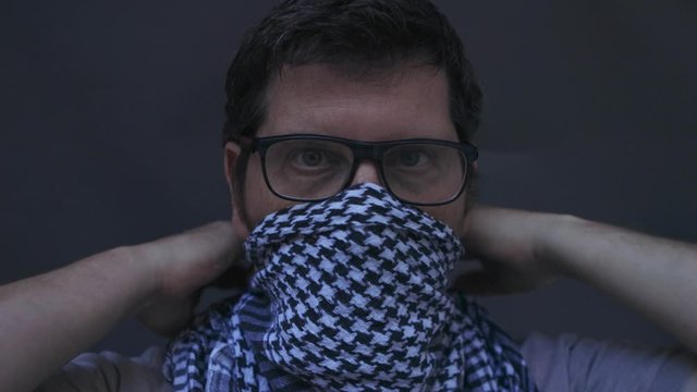 Person putting on checker motive scarf and securing it tightly under glasses and around the neck. Low key natural light studio shot against a dark grey background
