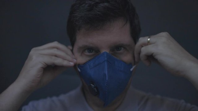 Closeup of caucasian male putting on and securing a dark blue n95 respirator with exhalation valve face mask. Low key studio footage normal speed -not slowed down- 60fps.