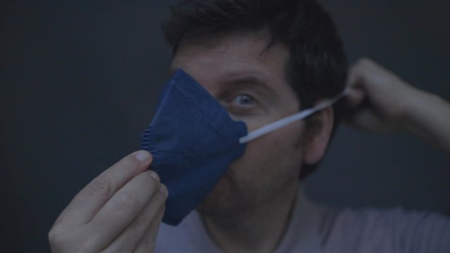 Caucasian male putting on and securing a dark blue n95 respirator face mask with exhalation valve. Low key studio footage slowed down from 60fps.