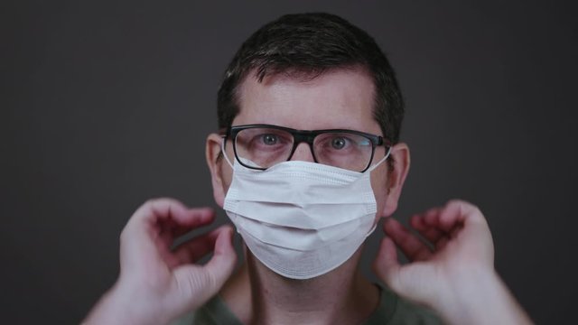 Caucasian male with blue eyes and glasses looking straight into the camera putting on a medical surgical face mask and securing it correctly covering mouth and nose against a grey background