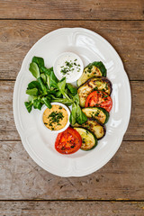 grilled zucchini, eggplant and tomato served on a white oval plate with spinach and sauces. on an old wooden brown table. view from above
