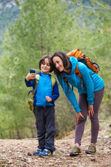 A child with a backpack takes a selfie on a smartphone with mom on the background of a mountain forest.