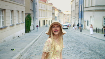 Cheerful pretty woman in white hat looking back at camera while walking in town.