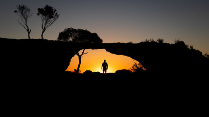 Silhouette of a man standing under a rock bridge at sunset