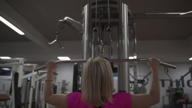 Fit blond women in pink shirt exercising medium grip lat pull down workout to strenghten up back muscles in gym. medium shot from behind