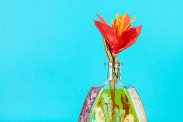 handmade artificial paper red flower in handpainted glass brandy bottle on aquamarine pastel color background