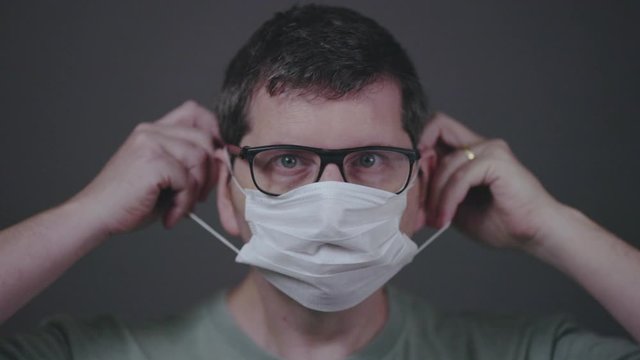 Caucasian male looking straight into the camera putting a surgical medical face mask on, securing it correctly covering mouth and nose. Low key studio footage slowed down from 60fps.
