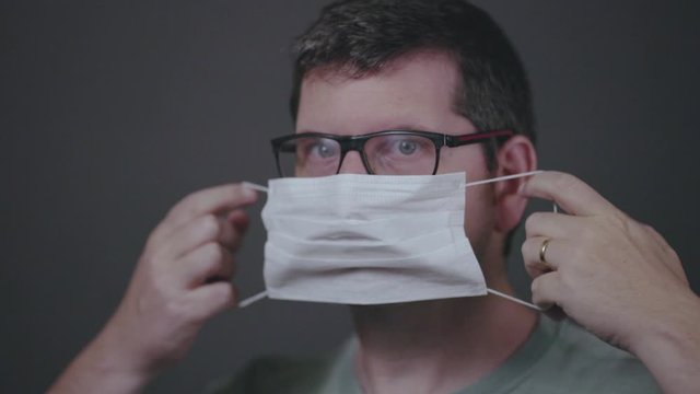 Sideview of putting on a surgical medical face mask, securing it correctly covering mouth and nose. Low key studio footage of caucasian male looking straight into camera slowed down from 60fps.