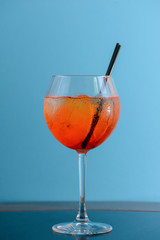 Cold summer alcohol cocktail with ice and orange in a wine glass and black plastic straw on light pastel blue background
