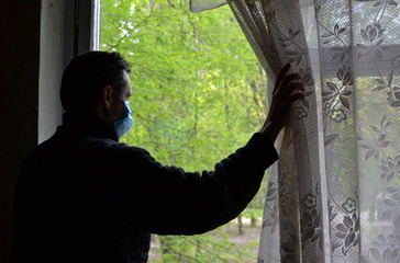 lonely sad man on self-isolation in blue surgical antiviral mask pushing back the curtain looks out...